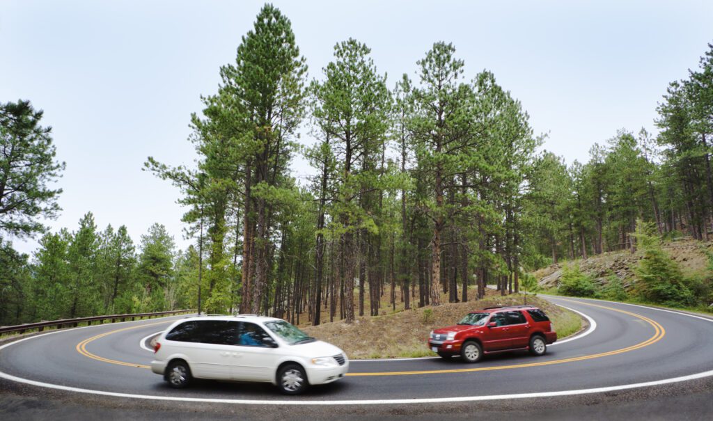 Tourists driving a red car and white van around a hairpin curve on a winding road through a forest